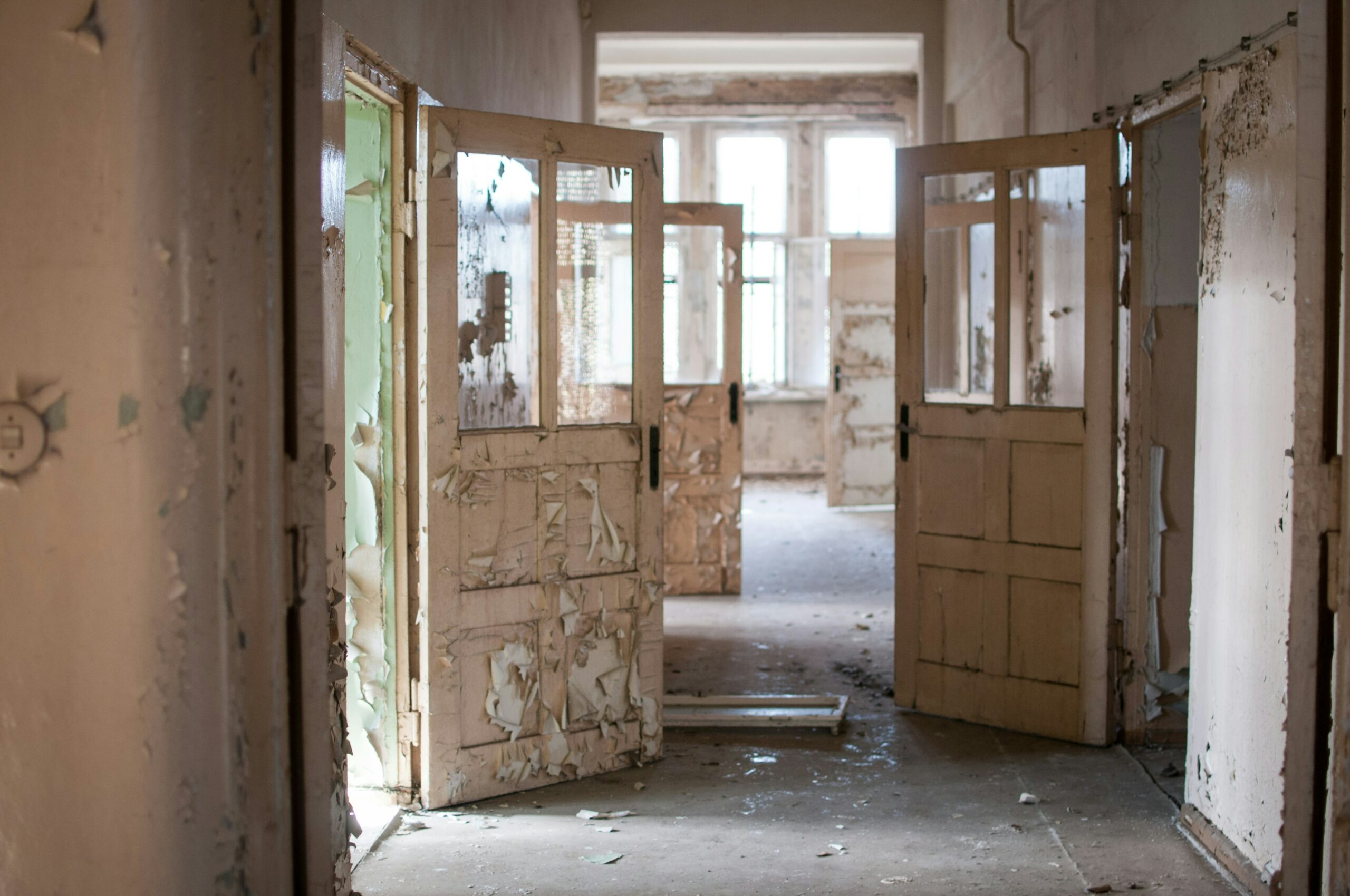 An empty hallway in an abandoned building has a series of open doors, each with peeling paint. Walking down this hallway is a journey.