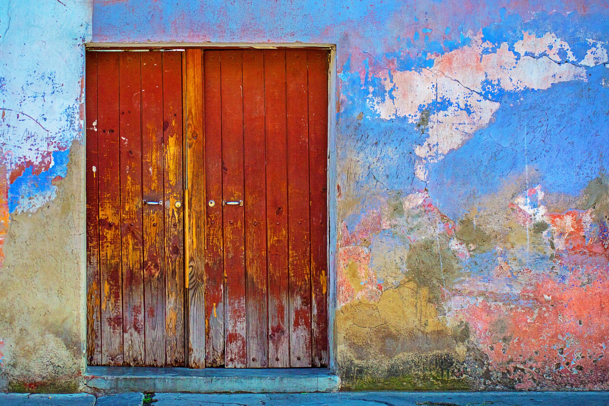 A two-panel cayenne door stands closed in the middle of a colorful wall with chipped and fading paint. Surely this wall has stories to tell about its community.